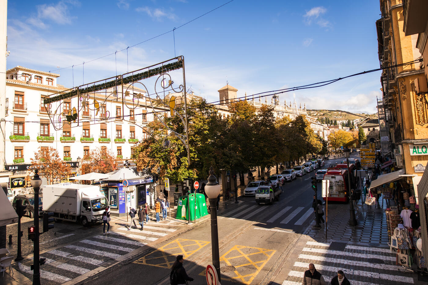 Calle del centro de Granada desde el balcón de la Suite Plaza Nueva: pasos de cebra, un camión de reparto parado, un microbús rojo y, al fondo, la fachada con pináculos de la Real Chancillería y el cerro del Albaicín.